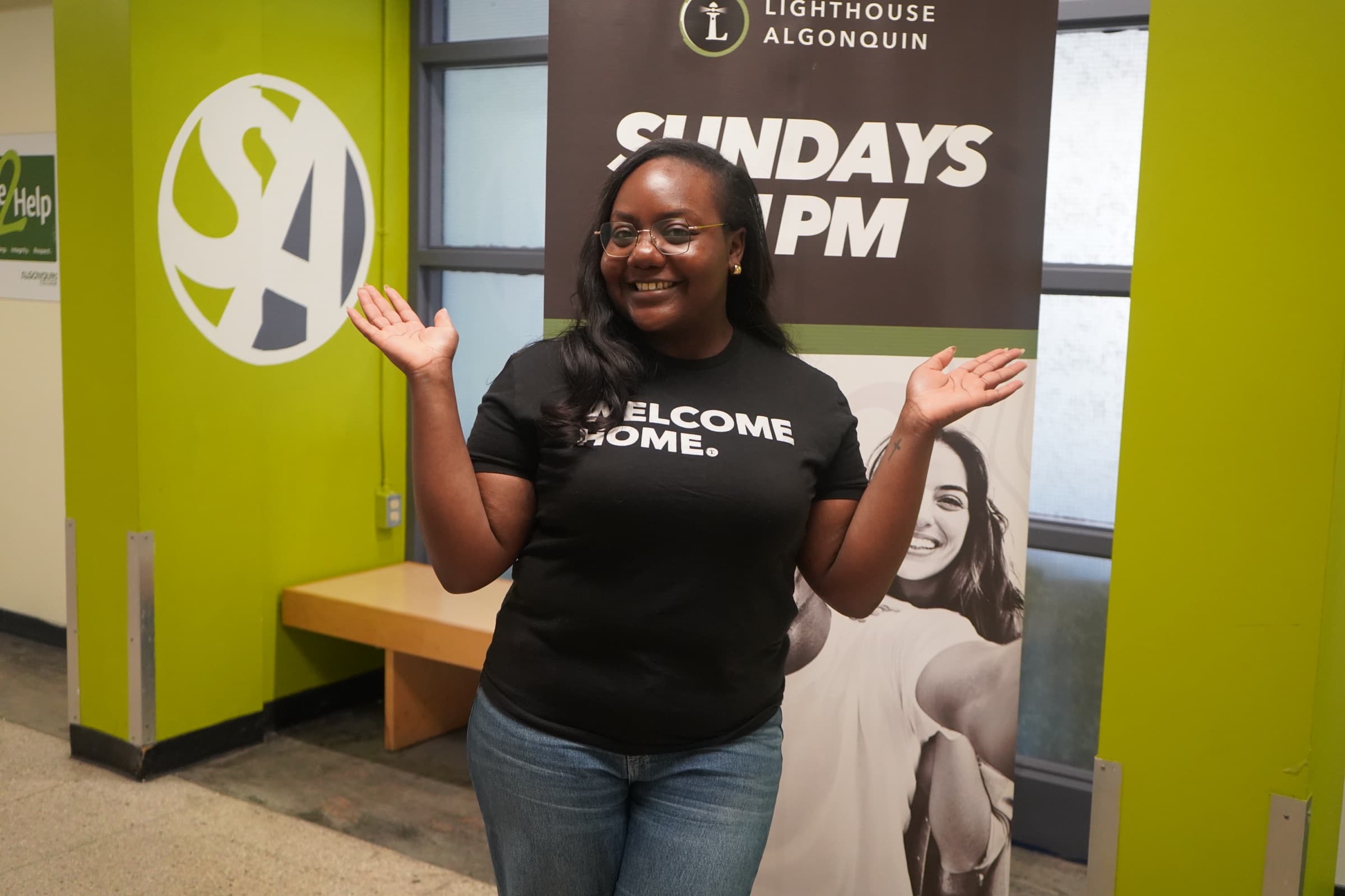 A greeter in a Welcome Home t-shirt, arms open in the hallway beside the Lighthouse Algonquin banner that reads Sundays 1 PM.