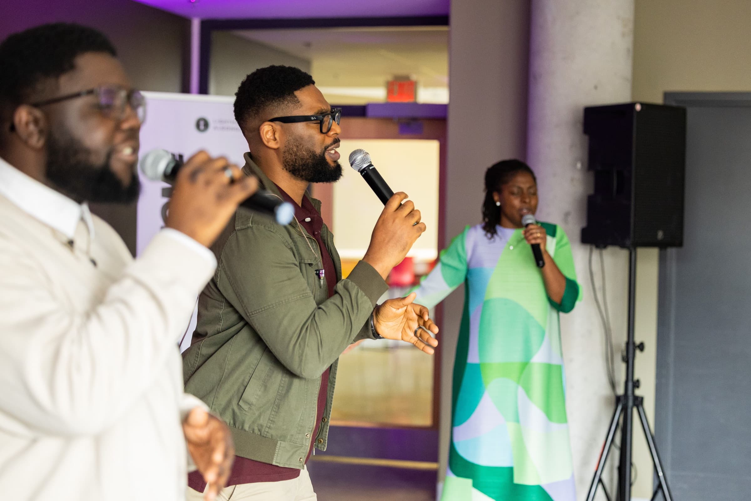 Three worship leaders singing into handheld microphones at the front of the room, the Lighthouse Algonquin banner behind them.