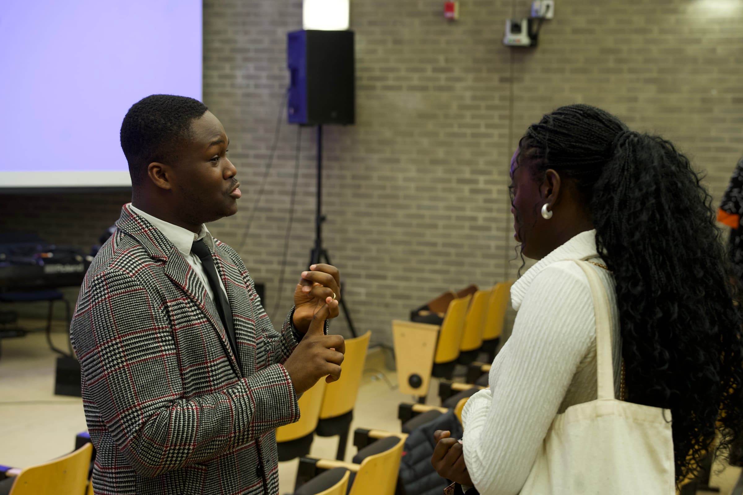 Two people talking in the lecture-hall seats after the service, one in a checked blazer mid-sentence and the other listening.
