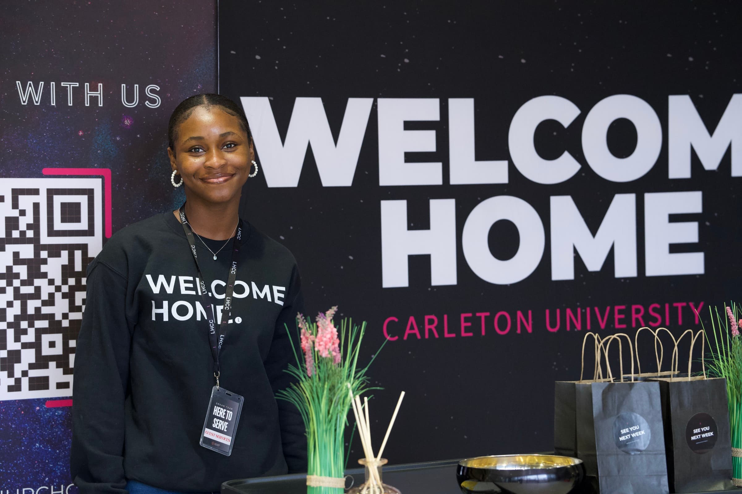 A Guest Services team member in a Welcome Home sweatshirt and lanyard smiling at the welcome desk, in front of a Welcome Home, Carleton University banner.