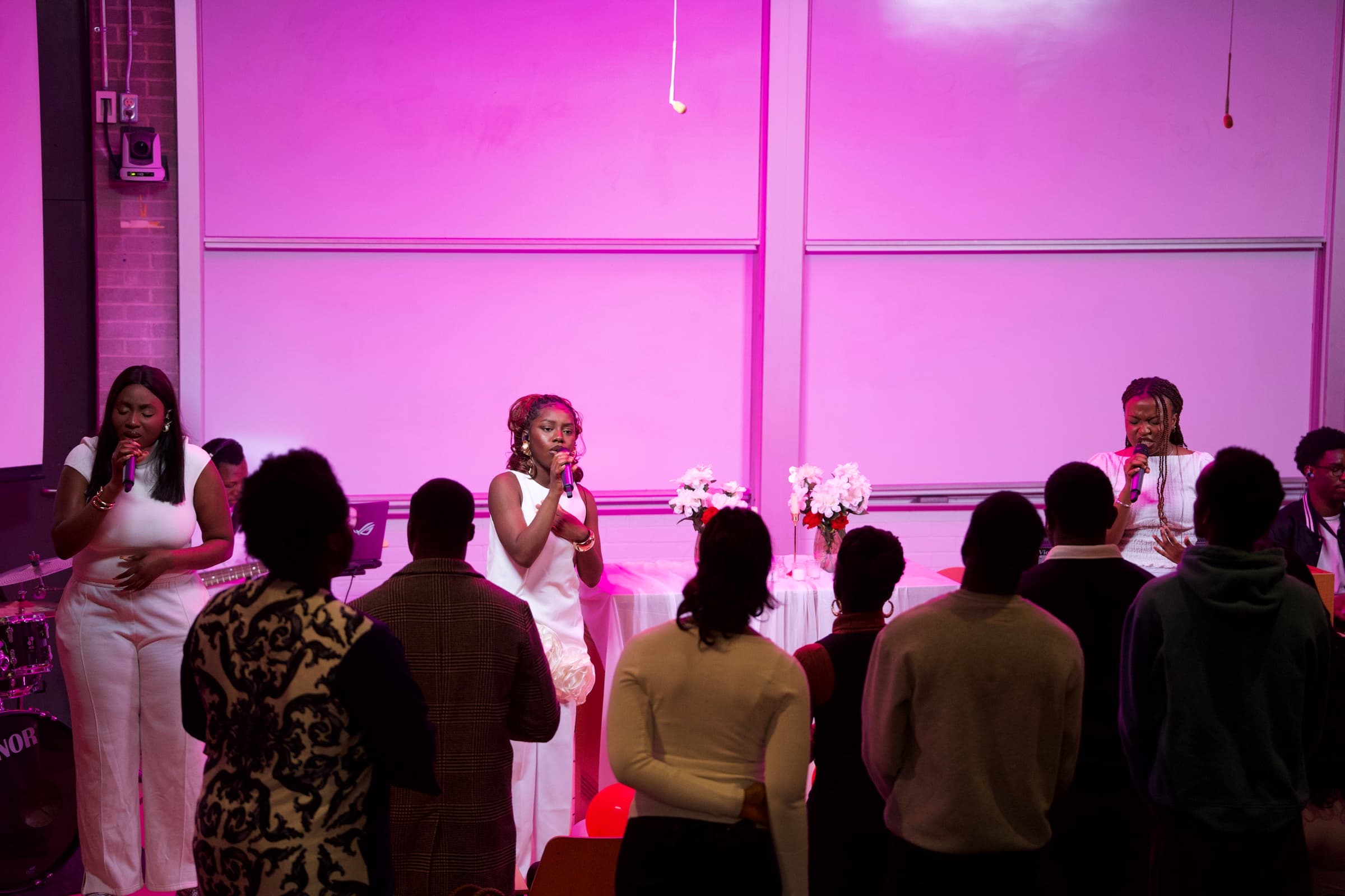 Three worship leaders in white singing at the front of a lecture room lit pink, the congregation standing with them.