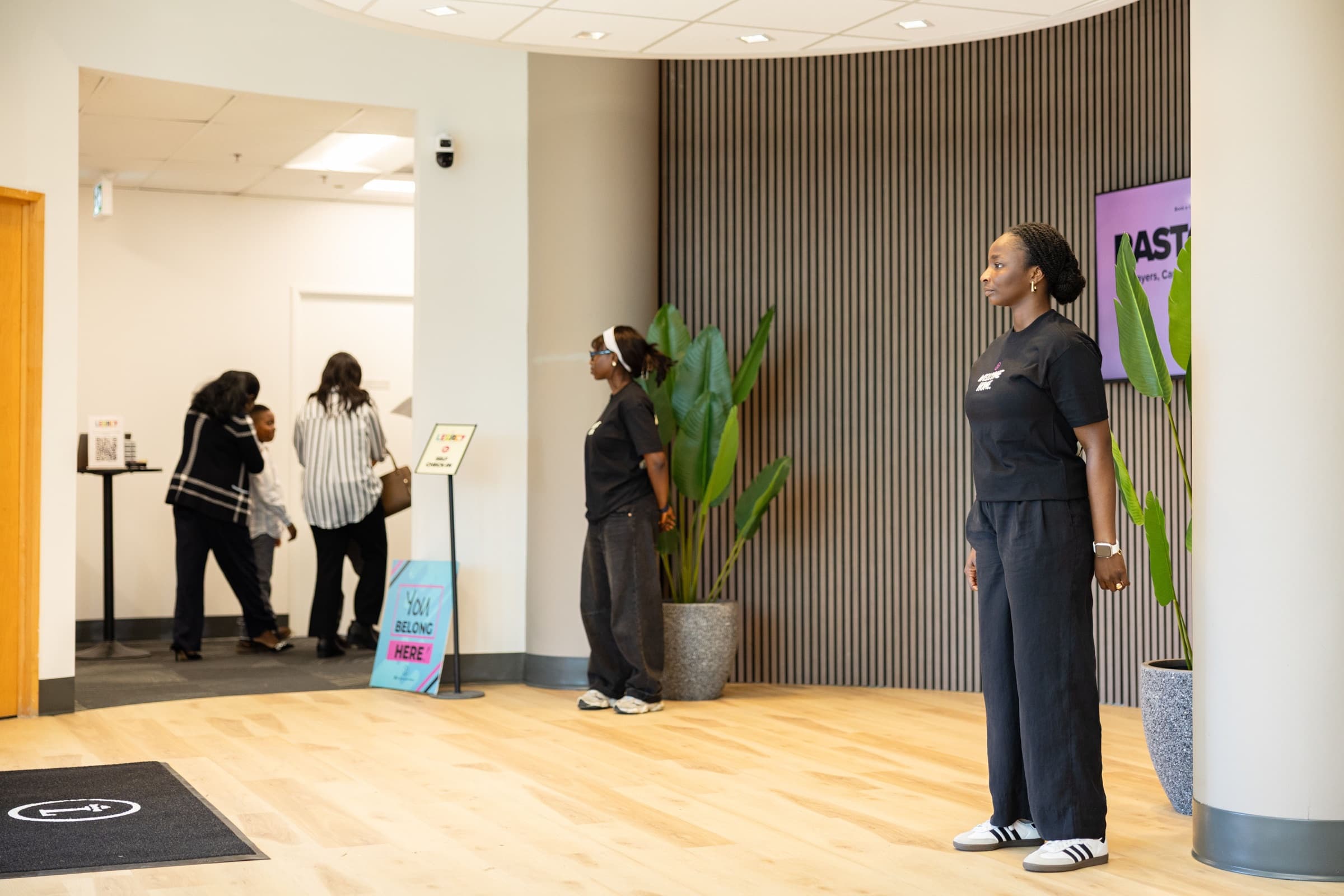 The Queensview lobby before a Sunday service, with a Guest Experience volunteer waiting by the doors, a family checking in at the kids desk, and a sign reading You Belong Here.