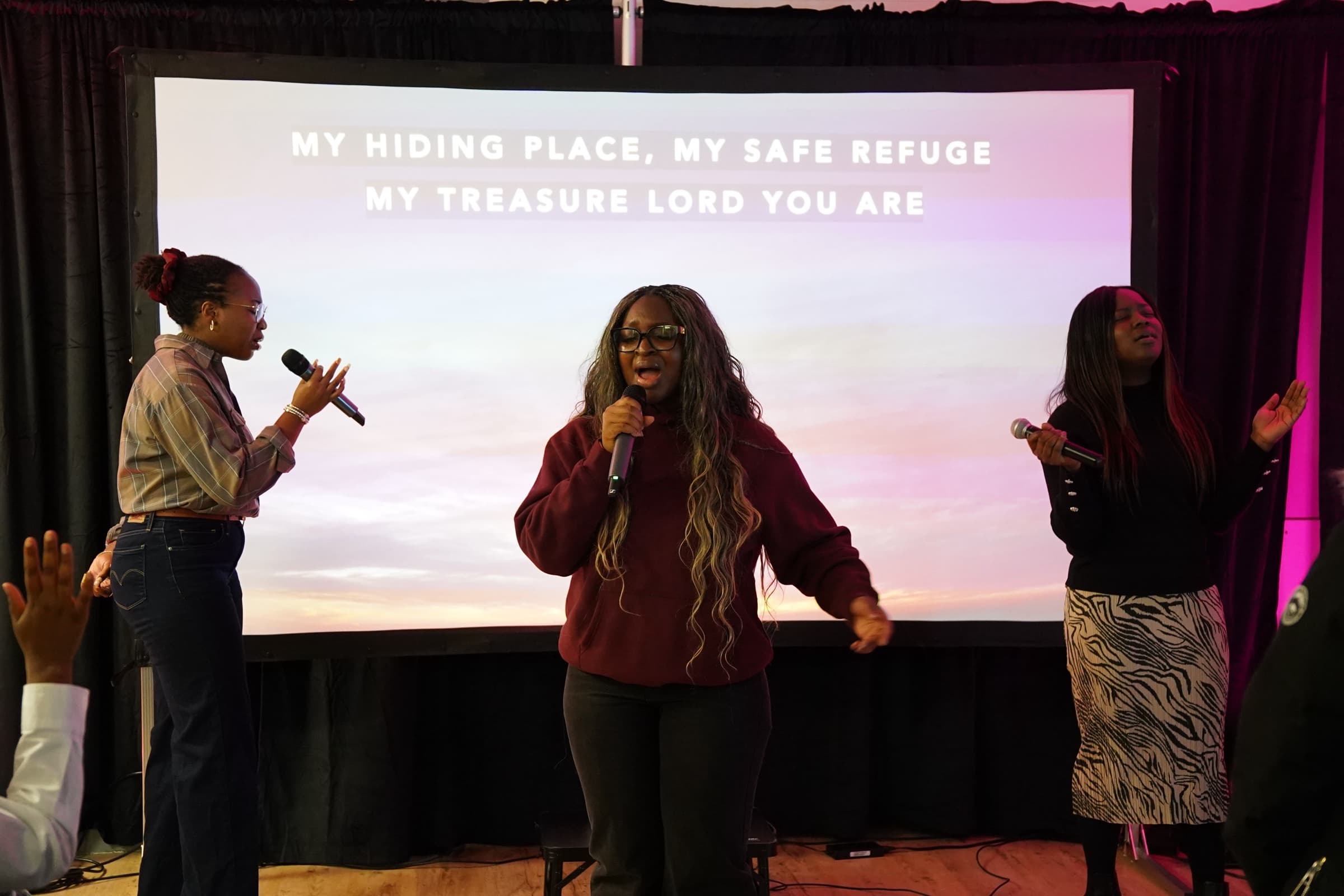 Three worship leaders singing into handheld microphones in front of a screen that reads My hiding place, my safe refuge, my treasure Lord you are.